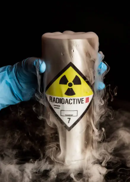A metal canister with a radioactive symbol sticker on it on a black background with smoke billowing out of it held by a hand wearing a blue protective rubber glove.
