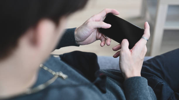 istock Close up view of young man holding horizontal mobile phone with blank screen while lying on couch at home. 1304832247