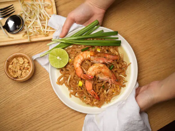 Cropped shot  of woman holding Pad Thai dish, stir fired Thai noodles with shrimps serving with lime, beansprouts and chives
