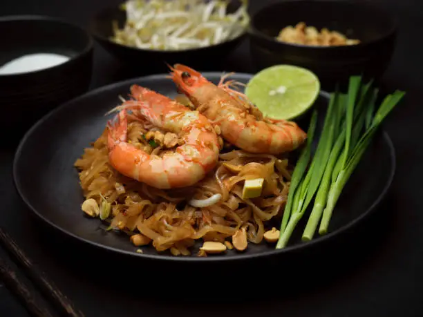 Close-up view of stir fired Thai noodles with shrimps serving on black plate with lime, beansprouts and chives, Pad Thai, black table