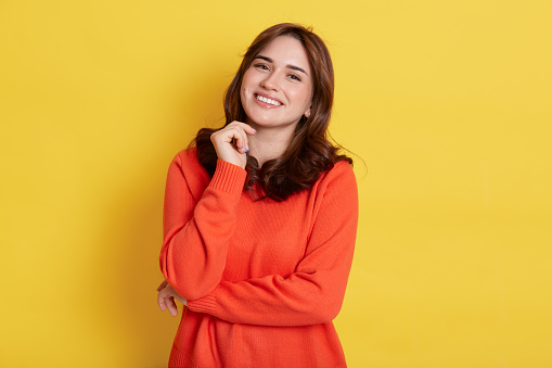 Happy smiling dark haired woman with pleasant appearance, wearing casually, looking at camera with toothy smile, keeping fist under chin, standing isolated over yellow background.