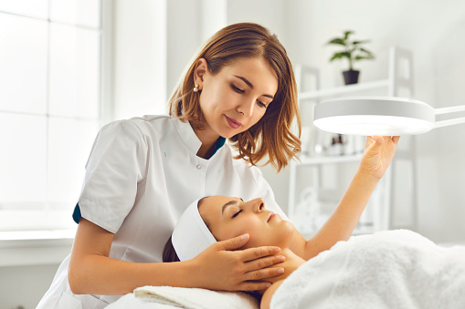 Young woman cosmetologist or dermatologist directing lamp for facial treatment to lying young woman in beauty spa salon. Facial treatment, massage, skincare, cosmetology concept