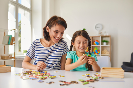 Young smiling mother with her little preschooler daughter make up puzzles while sitting at a table in the room. Concept of childhood, games and leisure.