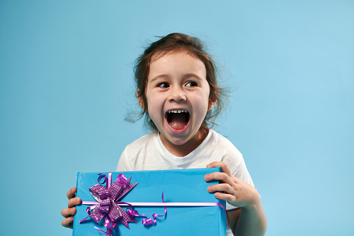 Adorable little girl laughing while posing on blue background with gift in her hands. Copy space. Celebration concept.