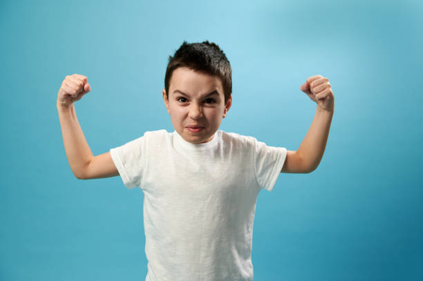 Boy pumping biceps and looking angry to camera on blue background with copy space