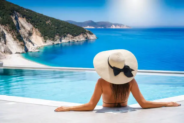 Photo of A woman with hat sits in the swimming pool and enjoys the view to the turquoise sea of Kefalonia island