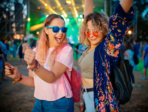 Two young women dancing and raising arms with joy on outdoor music festival, blurred background for generic look