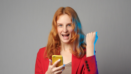 Young caucasian redhead woman in red jacket use phone rejoice isolated on grey copyspace