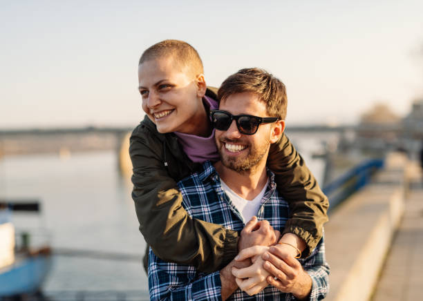Cancer cured woman and her partner walking near the river bank on sunny day Day in a life of cancer survivor - Mid-adult Caucasian woman fought lymph nodes cancer and finished series of chemotherapies with visibly burnt skin around neck and head, falling hair and dry skin. free hug stock pictures, royalty-free photos & images