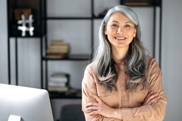 retrato de una exitosa mujer de pelo gris maduro, mujer de negocios, ceo o tutora de negocios, de pie en la oficina con los brazos cruzados, mirando y sonriendo amablemente a la cámara - hermoso ama de casa fotografías e imágenes de stock