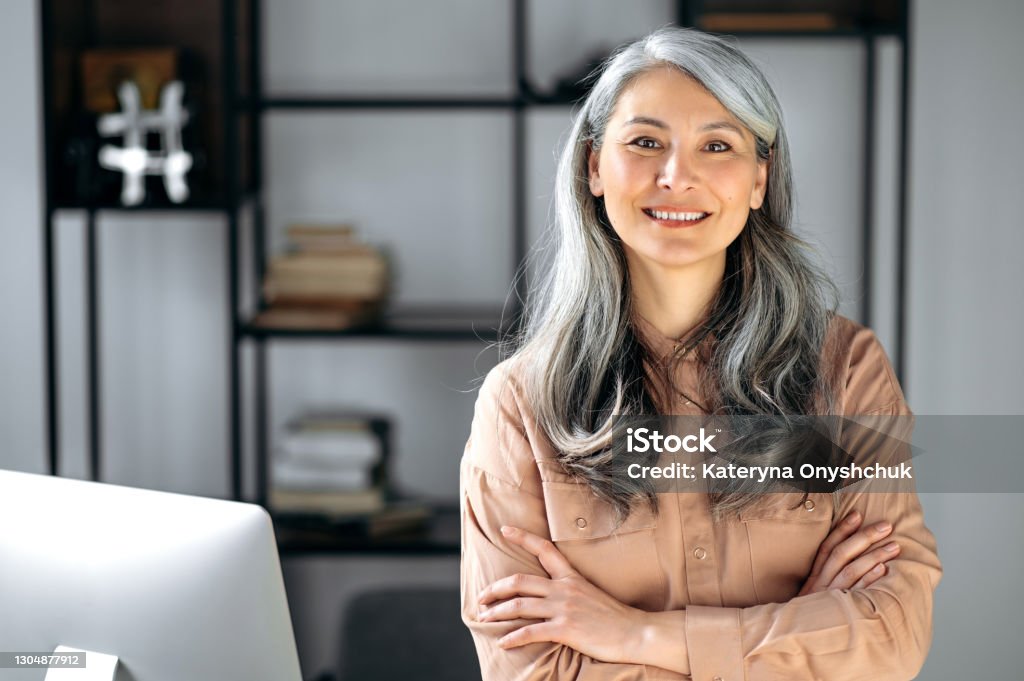 Portrait of a successful confident mature gray-haired lady, business woman, ceo or business tutor, standing in the office with arms crossed, looking and friendly smiling into the camera Women Stock Photo