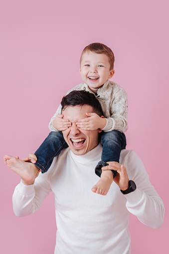 Thrilled happy euphoric boy riding on dad's shoulders, covering his eyes, blocking view. Over dark pink background.