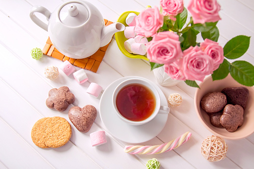 Setting the table for morning tea party. Сup of tea, teapot, bouquet of pink roses and sweets on white wooden table. Romantic breakfast