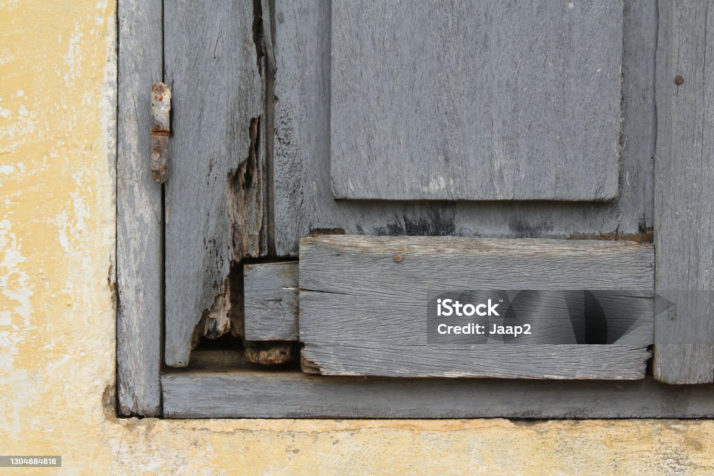 Wooden shutter on yellow African wall, weathered  and in bad condition Abstract front view close-up of a weathered wooden shutter with rusted nails and hinge, on a plastered wall painted in pastel yellow color Abstract Stock Photo