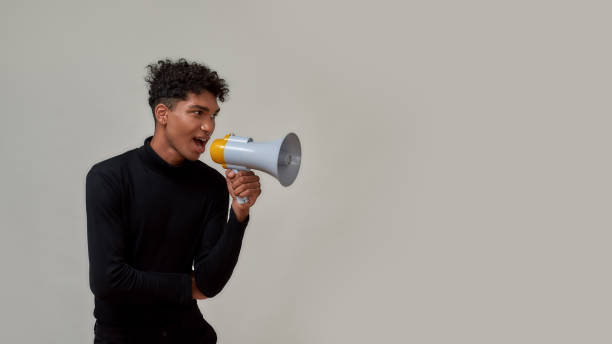 istock Cheerful young guy with piercing dressed in black looking aside and screaming in megaphone, making sale announcement, posing isolated over gray background 1304891067
