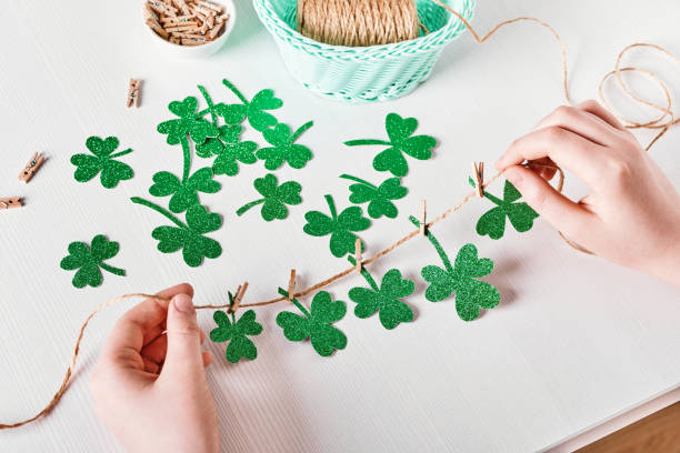 decoración diy st.patricks day. las manos hacen tréboles guirnalda de papel brillante en la mesa blanca - st patricks day irish culture child leprechaun fotografías e imágenes de stock