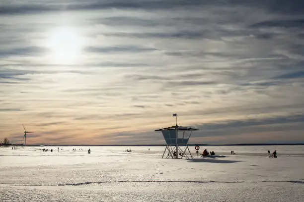 The life guard's hut is standing in guard while people are enjoying a beautiful winter day on the seaside. The sea is all frozen and the beach is covered in snow.