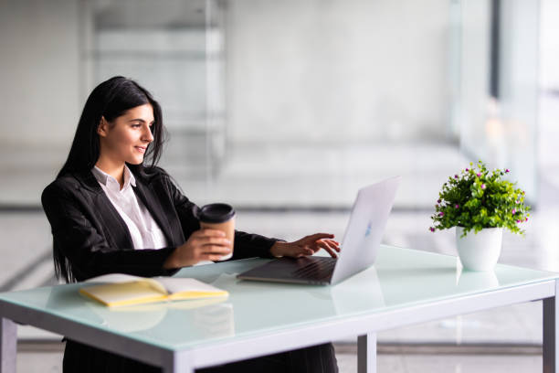 istock Attractive woman sitting at desk in office, working with laptop holding document, having takeaway coffee. 1304903019