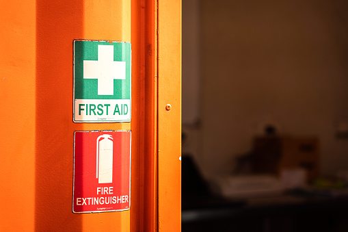 Close-up at first aid and fire extinguisher emergency equipment signs which is installed in front of the container medical room, with blurred background of inside the room (dark environment).