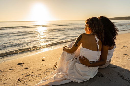 Two lesbian young women view from back sitting on seashore looking at sunset or dawn. Afro american homosexual girl hugs her blonde girlfriend near ocean water in summer vacations. Diversity concept