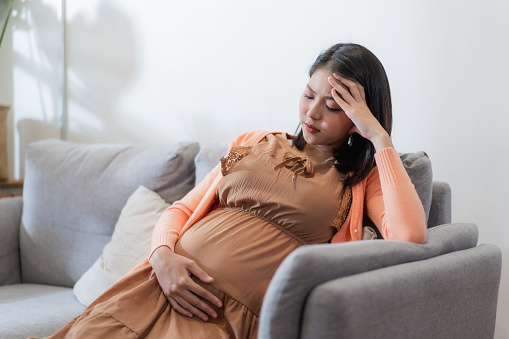 Young pregnant asian woman has suffering from headache sitting on sofa. Pregnancy symptoms
