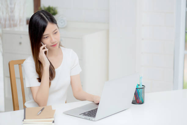 istock Young asian woman talking phone and work from home with laptop computer, freelance using notebook to internet online and speak on smartphone, domestic life, business and communication concept. 1304920513