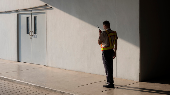 Asian security guard in safety vest walking on sidewalk and using walkie talkie or portable radio transmitter with sunlight and shadow on surface of staff room door in grey cement wall background