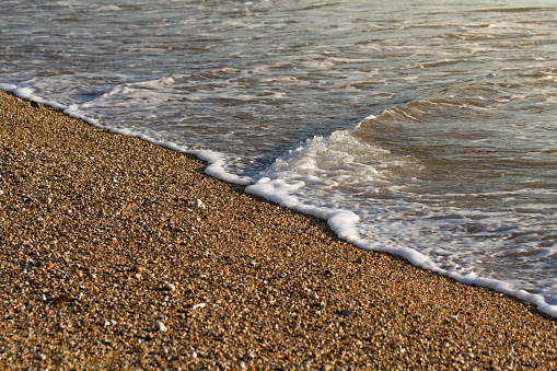 Close up of a pebble beach lit by a golden glow with gentle waves lapping at the water's edge in a tranquil seaside background