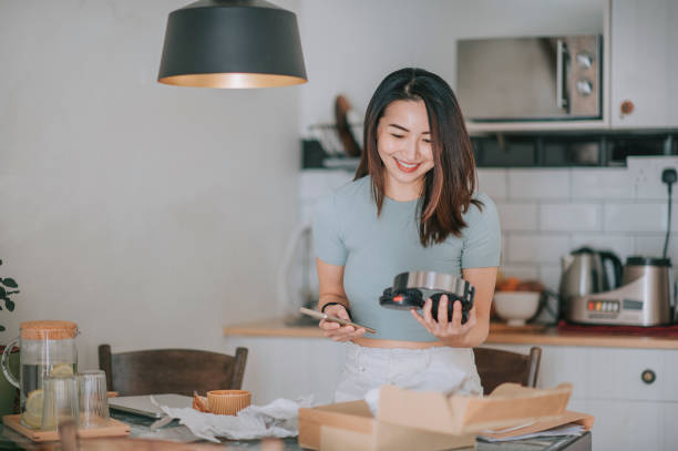 asiatico cinese bella donna che riceve disimballaggio cercando sulle sue cuffie dallo shopping online in cucina - unboxing foto e immagini stock