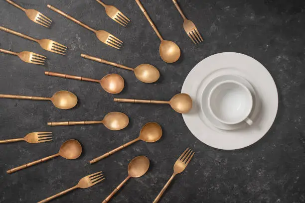 White ceramic plate, cup and brass forks and spoons look like sperm competition. Spermatozoons floating to ovule. Concept of fertilization, pregnancy and contraception. Top view, close up