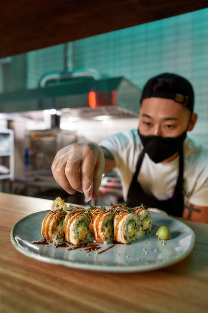 istock Professional sushi chef wearing protective mask and gloves decorating sushi rolls preparing to be served on plate at commercial kitchen 1304925648