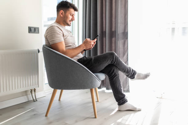 istock Handsome young man looking at his smart phone and smiling while sitting in big comfortable chair at home 1304925762