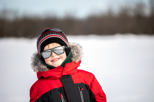 A cute redhead boy wearing sunglasses is walking outdoors with his family in the forest in winter after snowstorm. It is a beautiful sunny day. He is smiling and looking at the camera.