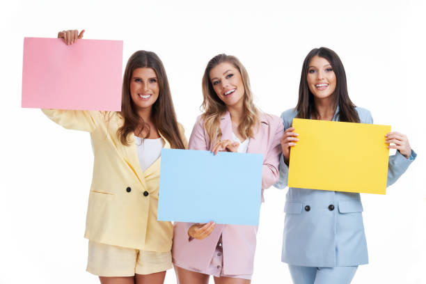 istock Three women in pastel suits holding billboard posing over white background 1304933008