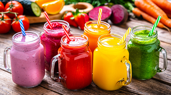 Healthy eating: fresh rainbow colored fruits and vegetables smoothies in Mason jars arranged side by side on rustic wooden table. Fruits and vegetables like tomatoes, cucumber, oranges, strawberries, beetroot, and carrots are out of focus at background. High resolution 42Mp studio digital capture taken with Sony A7rII and Sony FE 90mm f2.8 macro G OSS lens