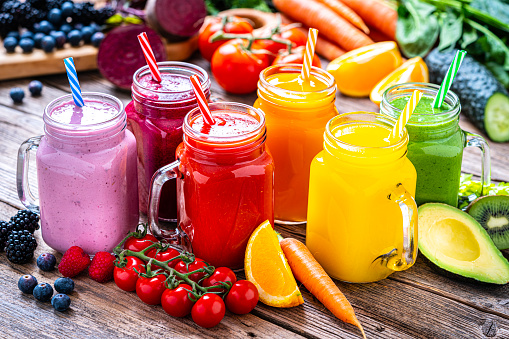 Healthy eating: fresh rainbow colored fruits and vegetables smoothies in Mason jars arranged side by side on rustic wooden table. Fruits and vegetables like tomatoes, cucumber, oranges, strawberries, beetroot, avocado and carrots are out of focus at background. High resolution 42Mp studio digital capture taken with Sony A7rII and Sony FE 90mm f2.8 macro G OSS lens