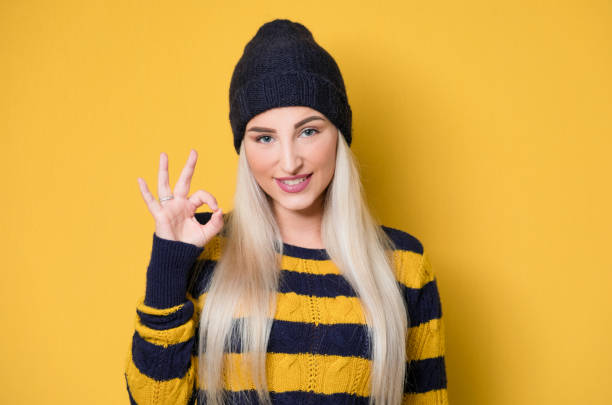 Joyful woman showing excellent sign with finger, model wearing woolen cap and sweater, isolated on yellow background. Yes or excellent gesturing, body language concept stock photo