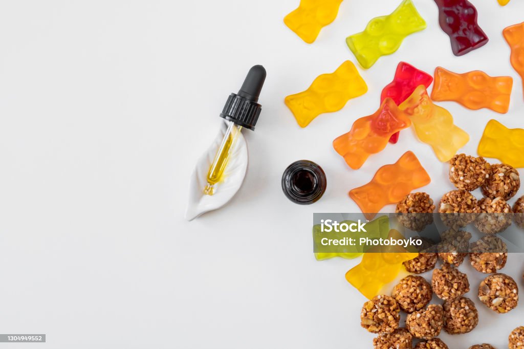 Gummy bears, cannabis cookies and cbd oil dropper in white backdrop. THD or cbd sweets in form of gelatine gummi bears and essential oil pipette background: calming edible food supplements Cannabidiol Stock Photo
