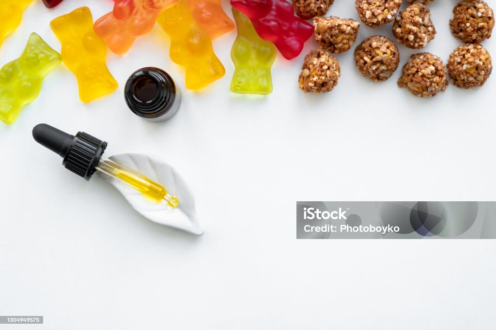 Gummy bears, cannabis cookies and cbd oil dropper in white backdrop. THD or cbd sweets in form of gelatine gummi bears and essential oil pipette background: calming edible food supplements Gummy Candy Stock Photo