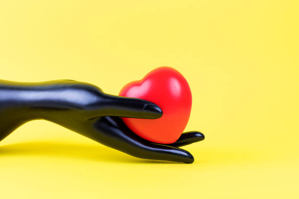 istock Mannequin women's black plastic hand holding plastic heart on a yellow background. 1304954726