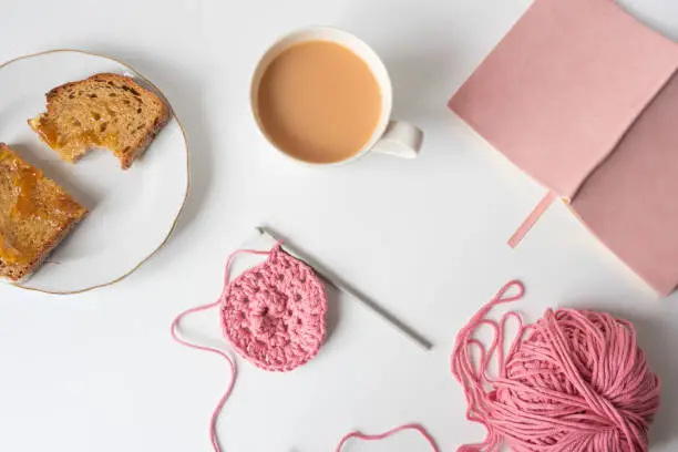 High angle view of self care objects on white table including cup of tea, pink crochet, toast on plate and journal
