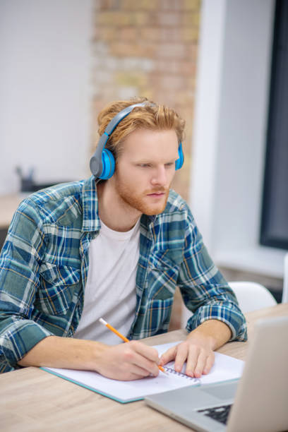 istock Ginger bearded guy in earphones sitting at the table and making notes 1304964754