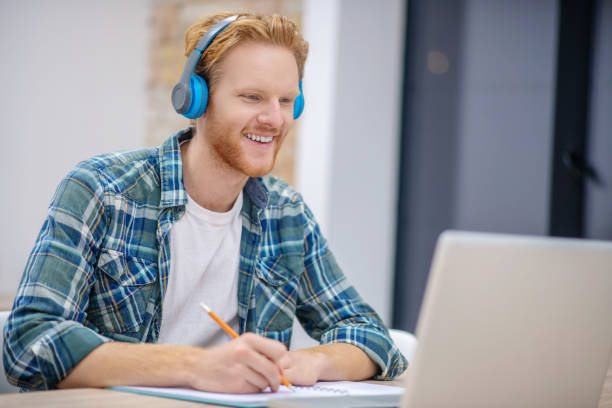 istock Ginger bearded guy in earphones sitting at the table and making notes 1304964755