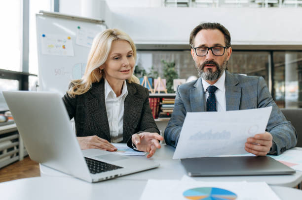 istock Concentrated mature business partners learning financial graphs for new project, doing paperwork. Elegant woman and serious man in formal suits sitting at the desk, look at the documents, teamwork concept 1304964866