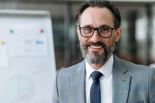 Close-up portrait of mature bearded businessman dressed in stylish suit and eyeglasses, looking at the camera with friendly smiling, standing in the office