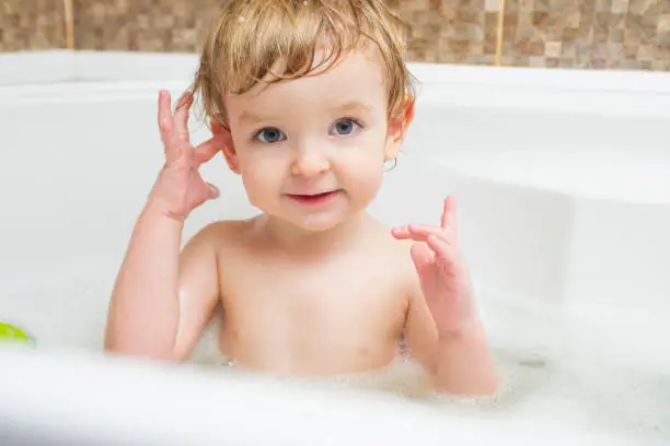 Photo of Funny little boy getting a bath in the bathroom. A child bathes in water with soap suds in white bath.