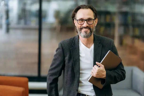 Portrait of happy confident businessman in eyaglasses and stylish formal suit. Middle aged successful male business leader looking directly at the camera, smiling, holding laptop in arms