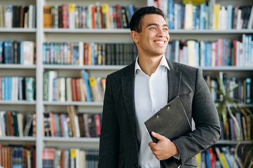 Happy successful young student or freelancer in stylish formal suit stands in modern office or library. Satisfied male employee looks away, smiling thinking about new project