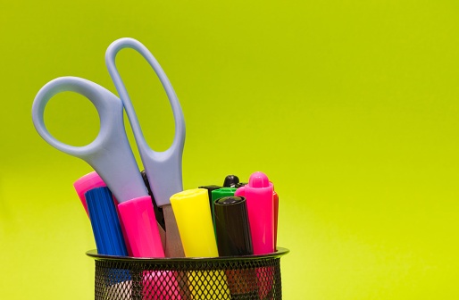 Mesh pen cup full of colorful markers, writing implements and scissors on a plain green background with copy space, concept image.