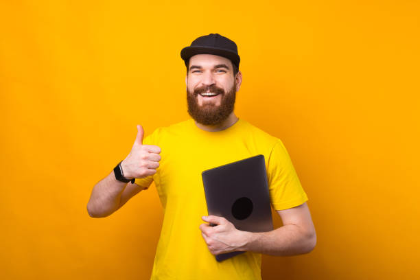 istock Smiling handsome bearded man showing thumb up and holding laptop 1304974560